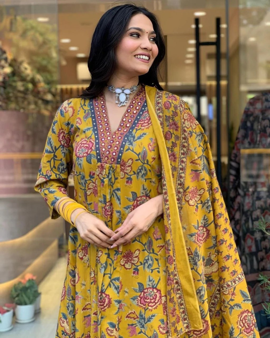 Woman wearing a colorful floral dress with a yellow dupatta in an indoor setting.